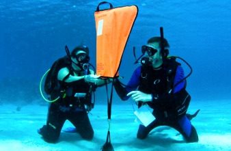 Two divers kneeling underwater demonstrating the deployment of a lift bag